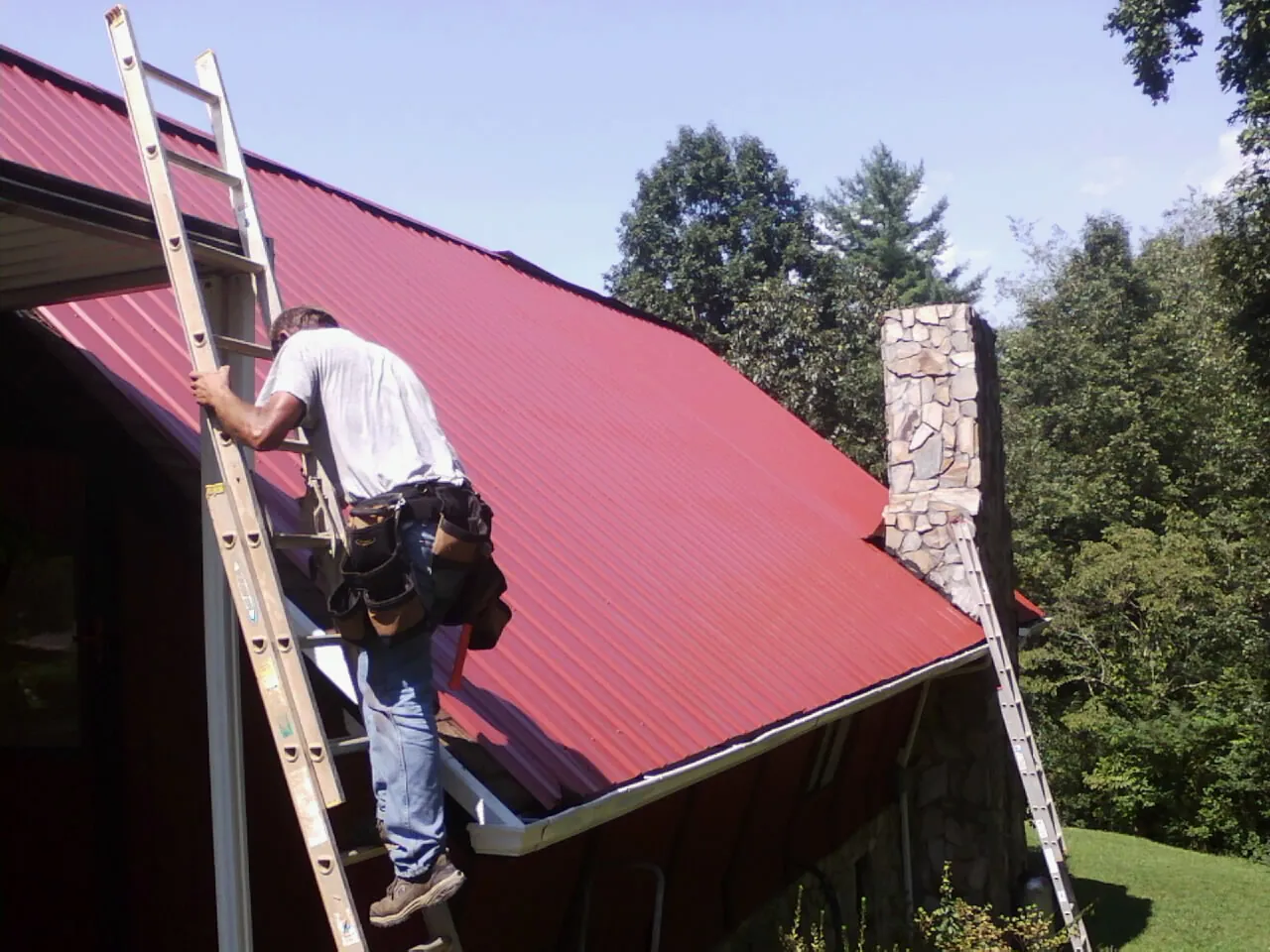 Roofer on a ladder beside a red metal roof and stone chimney — Soffit & Fascia Repair by Roofing Brothers in Anderson